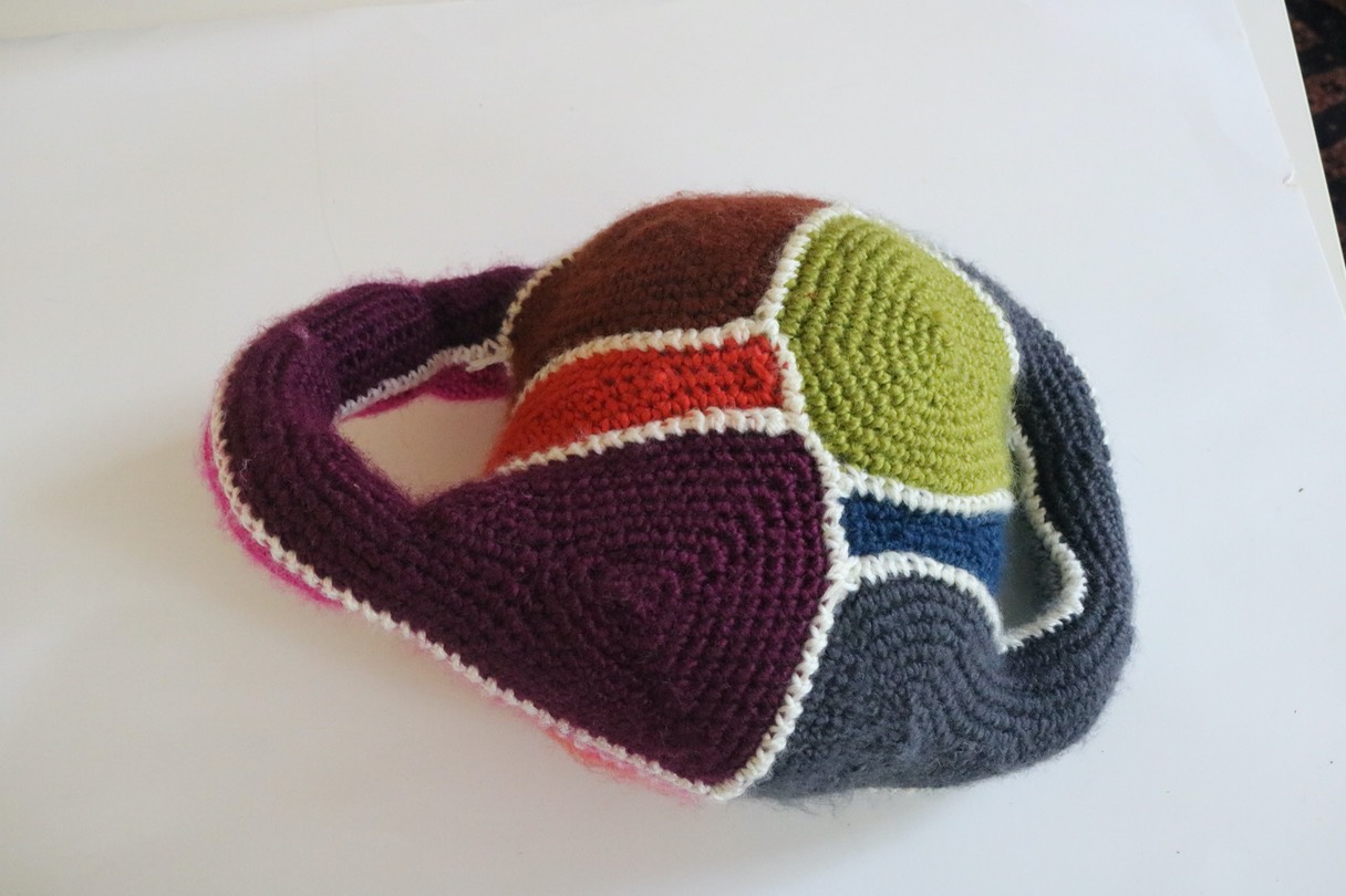 Picture of a crocheted baseball cap with sections of blue, green, gray, purple and orange joined by white stitching. 